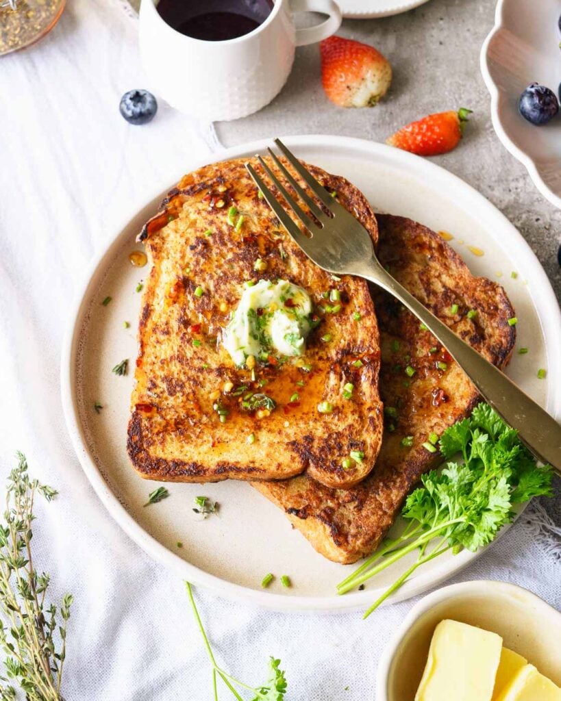 Savory french toast on a white plate, garnished with chives, herb butter and hot honey, with coffee on the side.