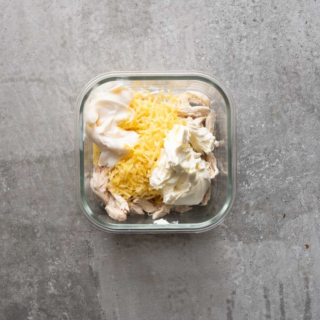 shredded chicken topped with Cream cheese, cheddar cheese in a square glass bowl.