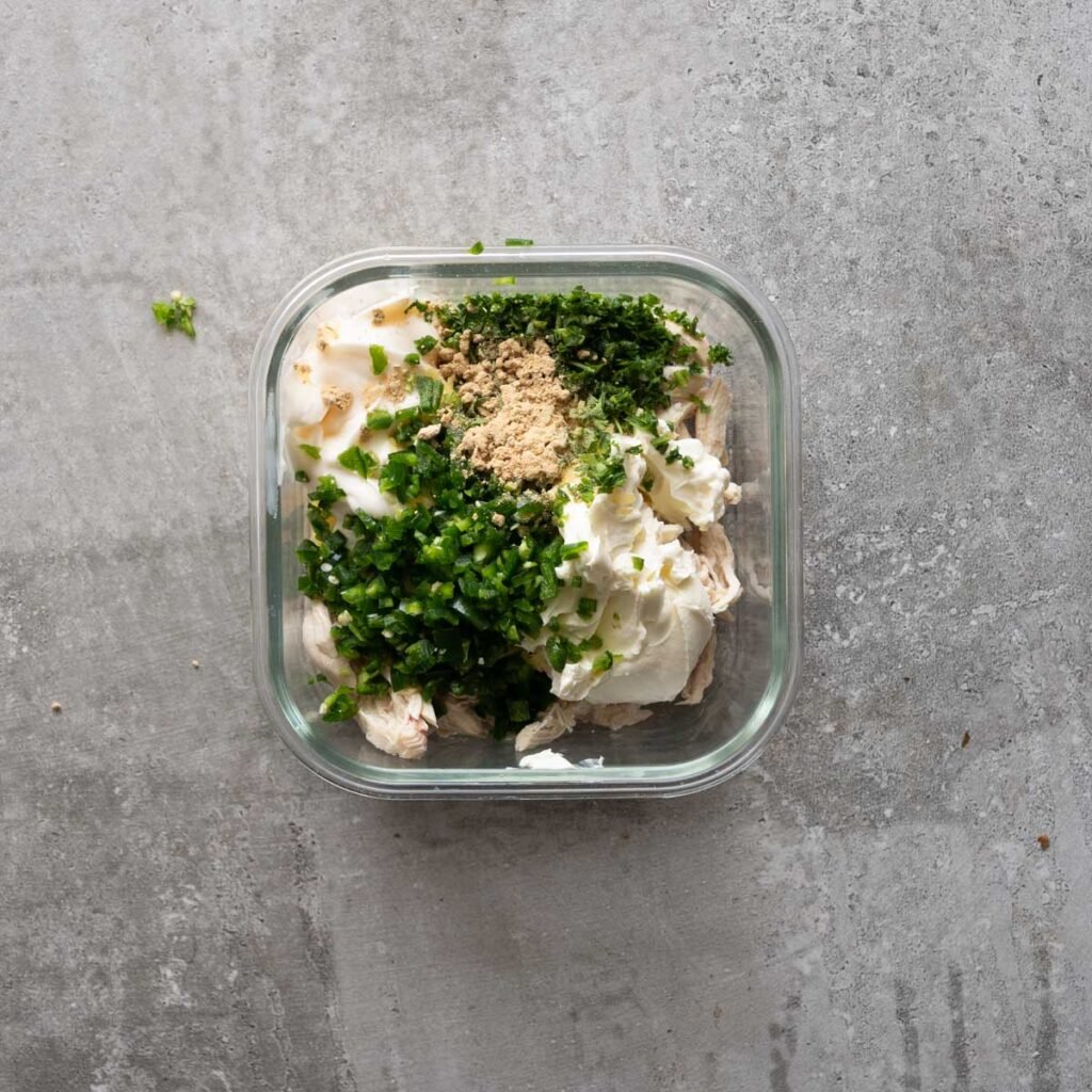 shredded chicken topped with Cream cheese, cheddar cheese , jalapeno, garlic powder and parsley in a square glass bowl.