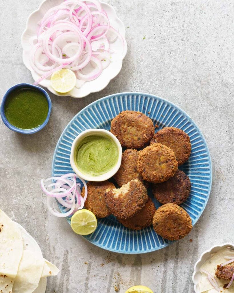 A blue plate with 10 shami kebabs, served with a green chutney, lemon and sliced onions.