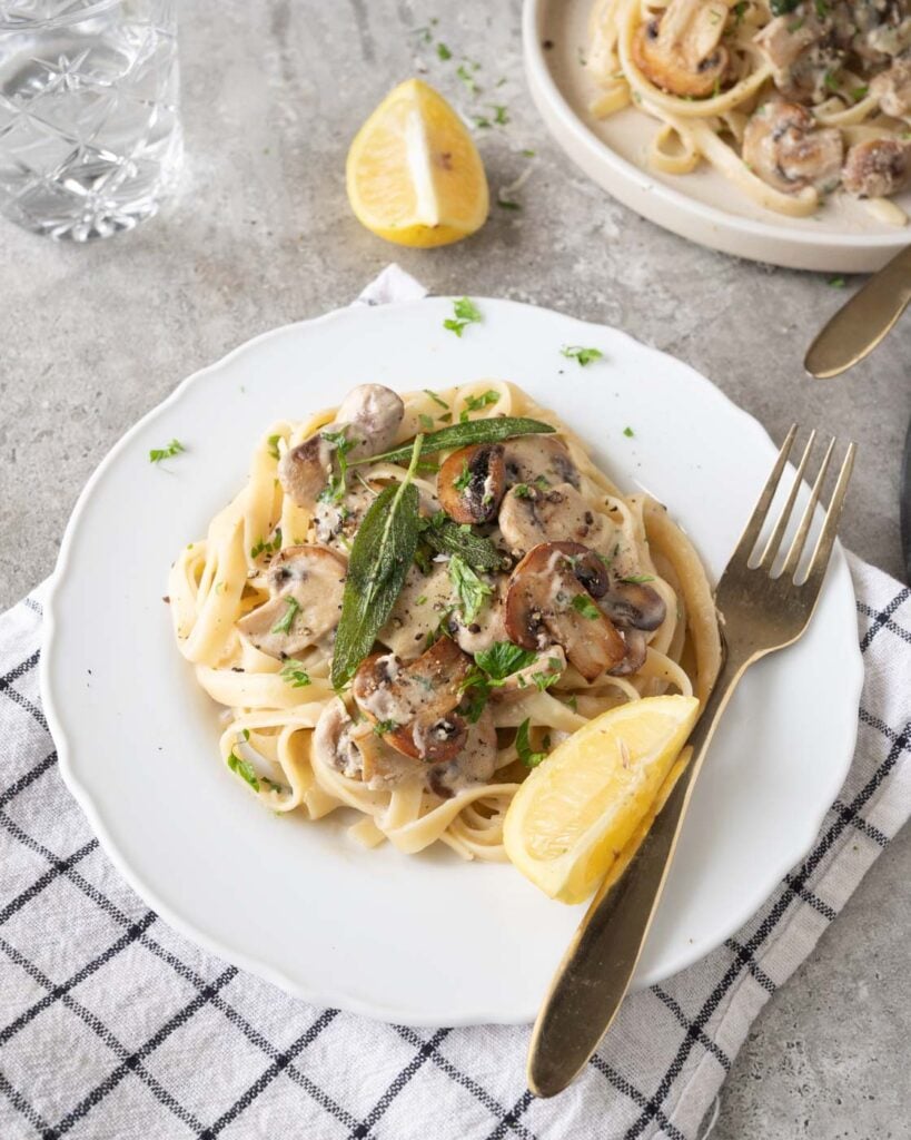 Truffle mushroom pasta on a white plate, garnished with caramalized golden mushroom, crispy sage leaves.SErved with a wedge of lemon