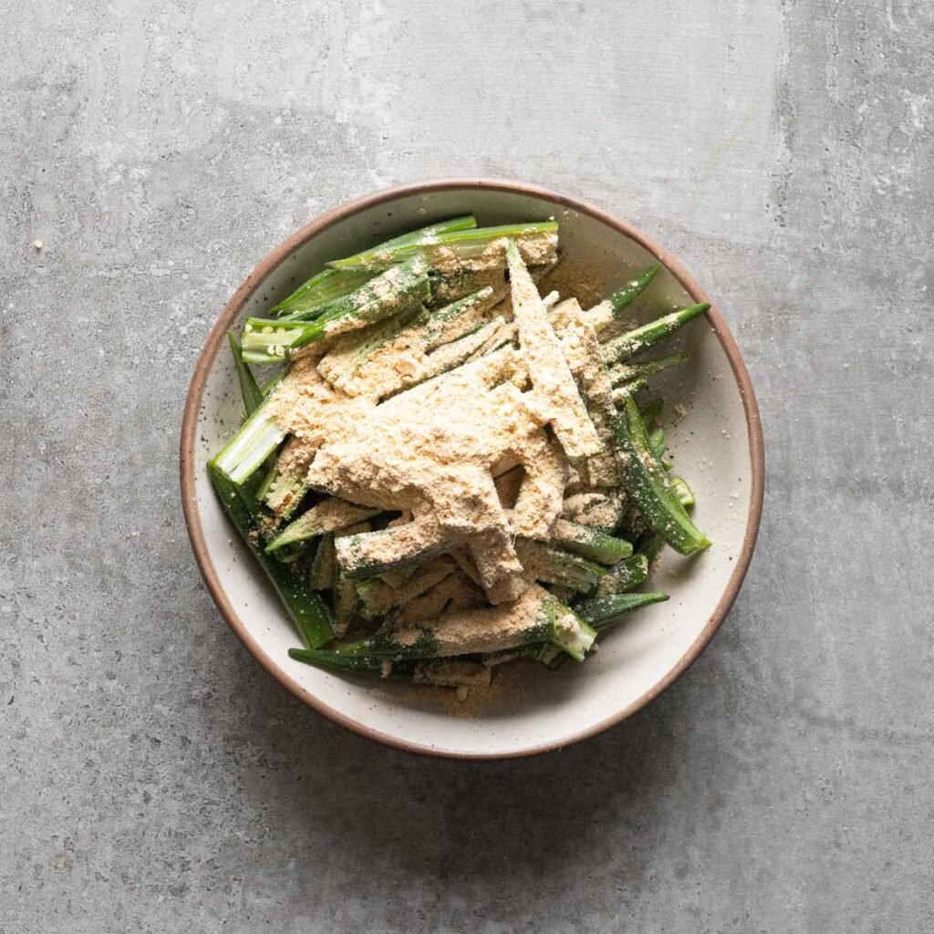 Okra cut into half, length wise with chickpea flour and spices on it, in a white bowl