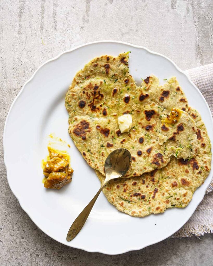 Zucchini paratha on a white plate with butter and pickle on the side.