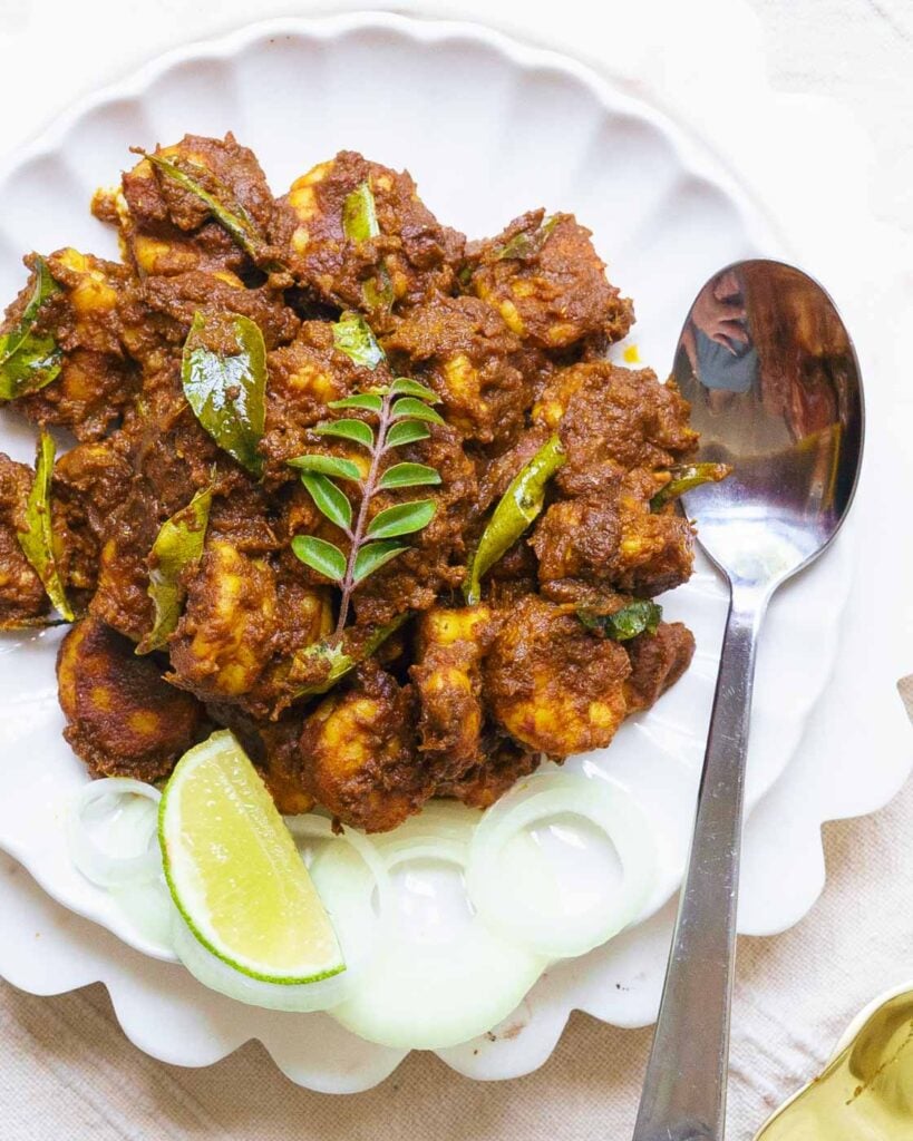 tawa masala shrimp also called prawn roast on a white fluted plate with a serving spoon next to it. Garnished with curry leaves